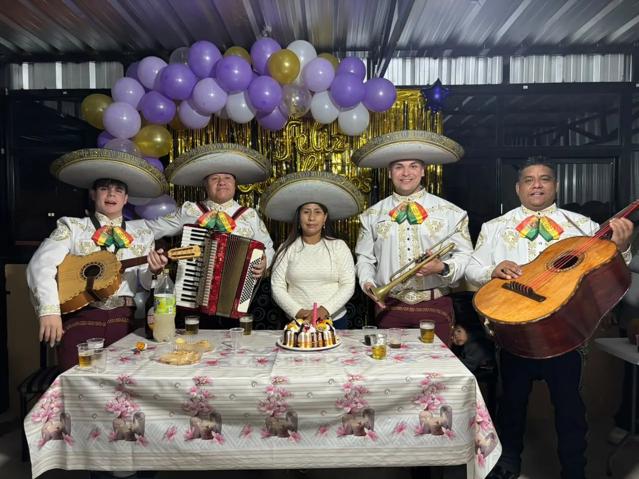 Cuatro mariachis con sombreros y moños tricolor, junto a una cumpleañera y su torta, en una fiesta con globos violetas y dorados