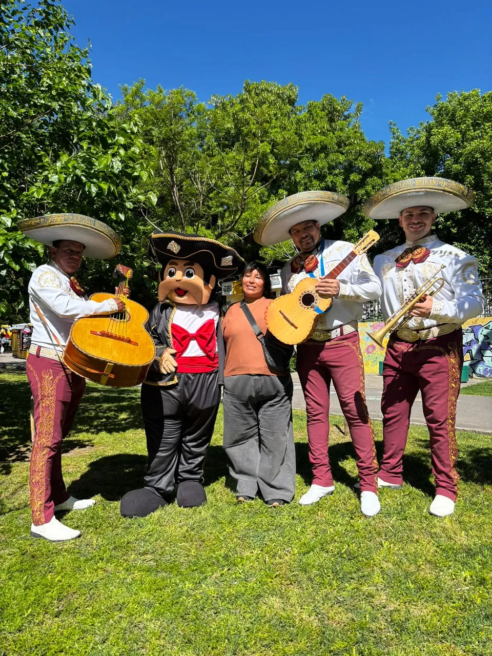 Cuatro mariachis y el muñeco Toribio posan junto a una mujer en un parque, un día de sol
